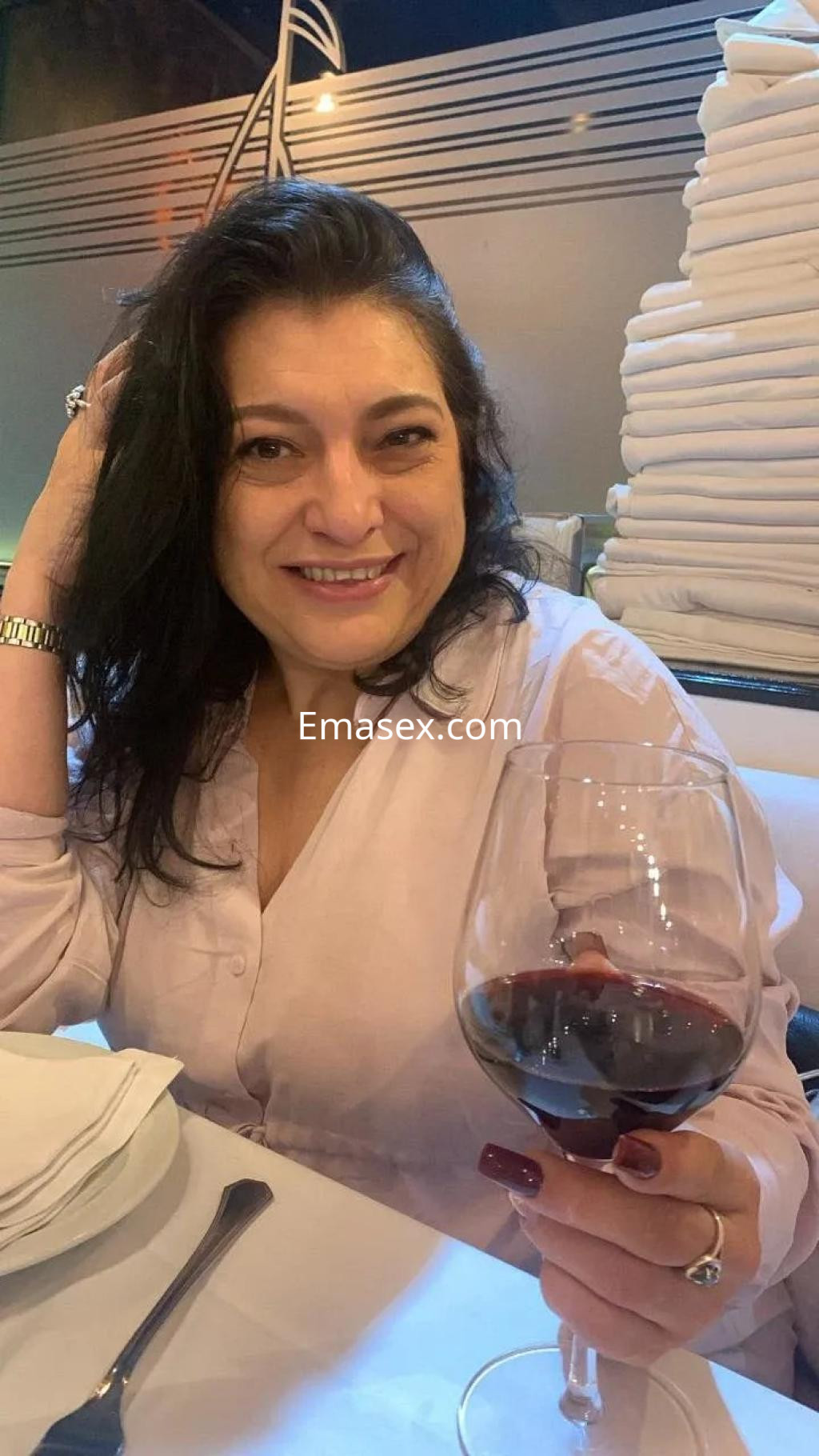 La puta en Alicante sonríe mientras sostiene una copa de vino en un ambiente elegante y bien iluminado.