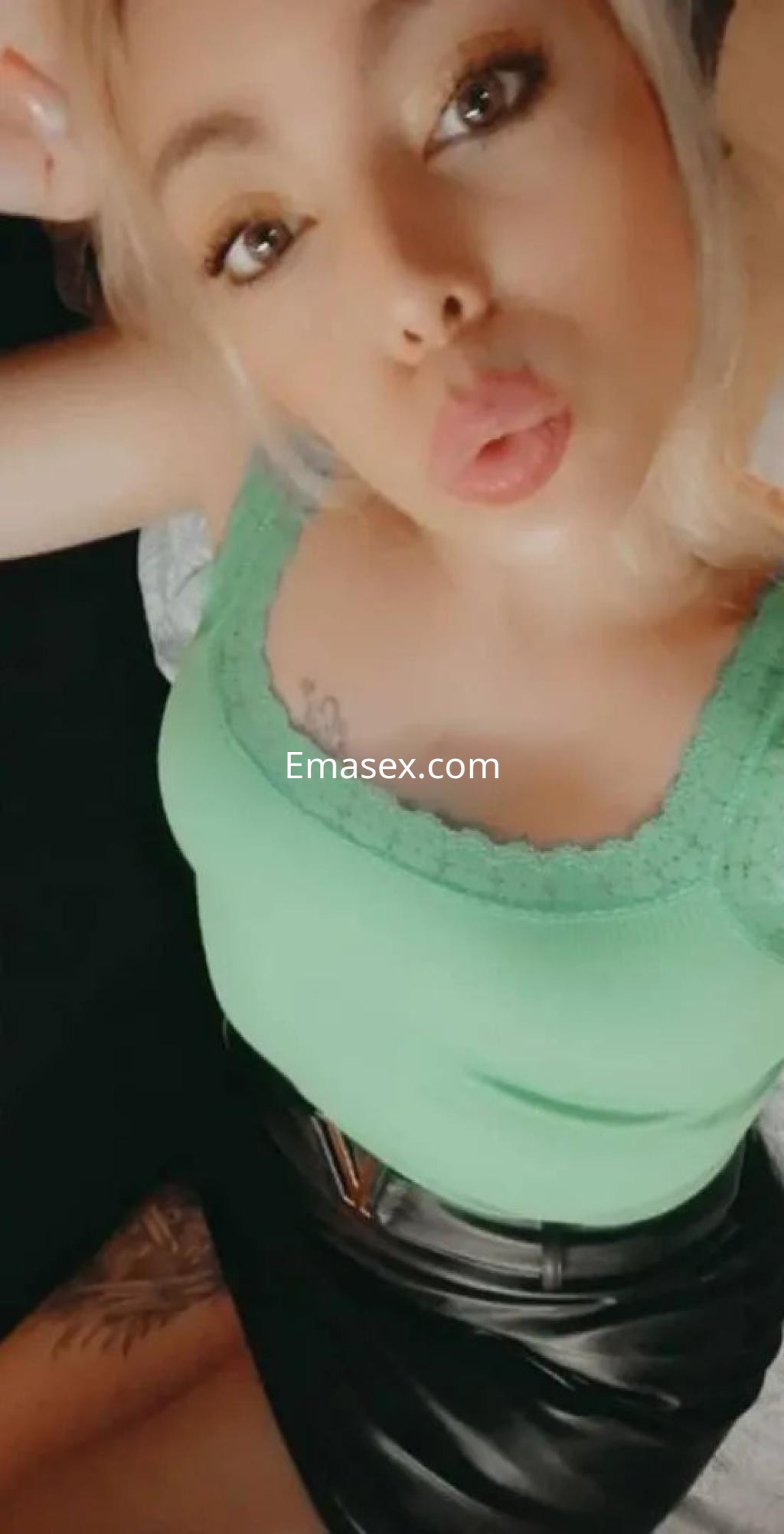 Puta en Alicante luciendo una blusa verde y una falda negra ajustada, posando con una expresión coqueta.