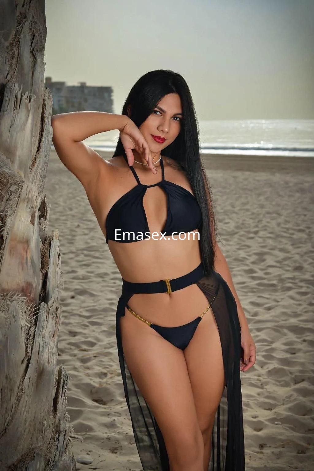 Escort de cabello largo y oscuro, luciendo un bikini negro con detalles dorados, posando en la playa de Alicante.