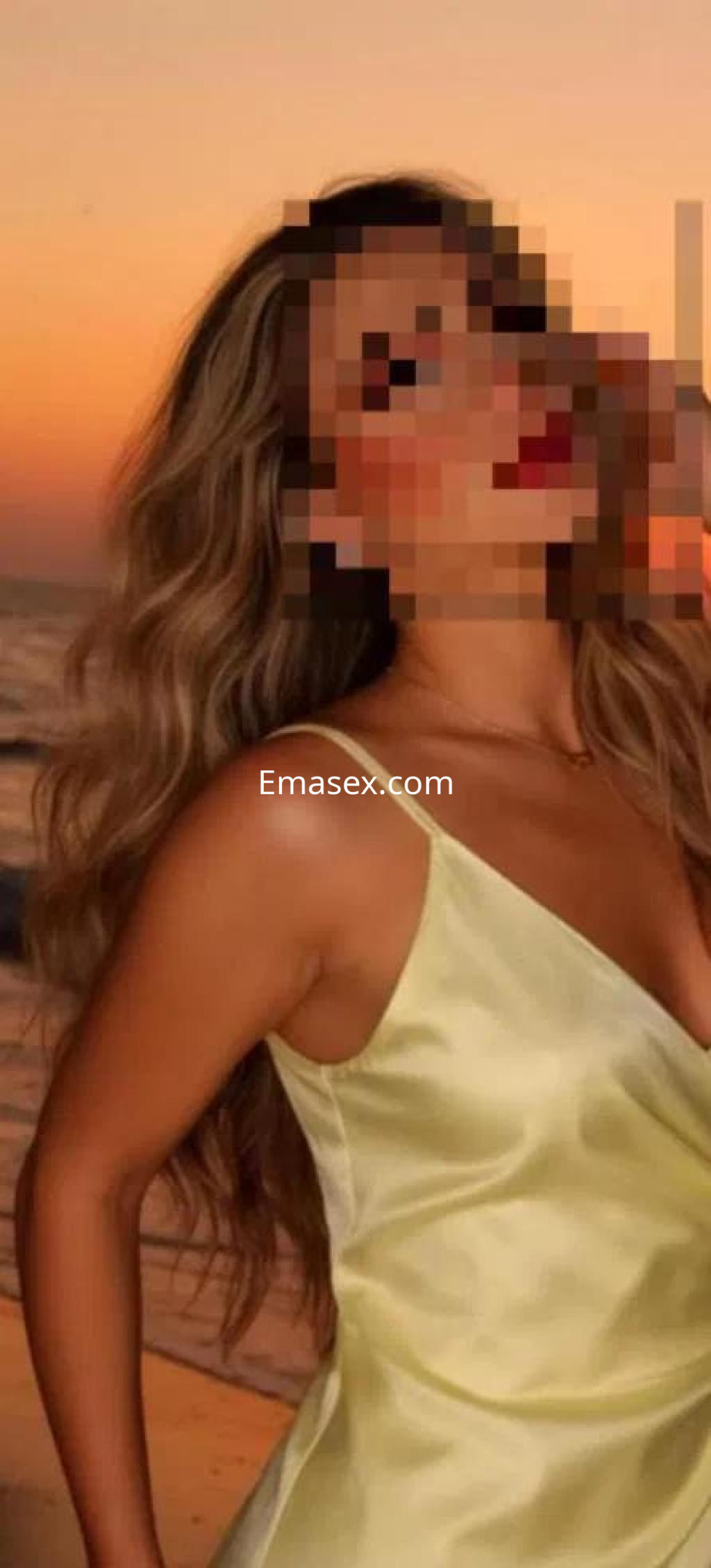 Puta de Badajoz con cabello largo y ondulado, viste un vestido amarillo y posa al atardecer junto al mar.