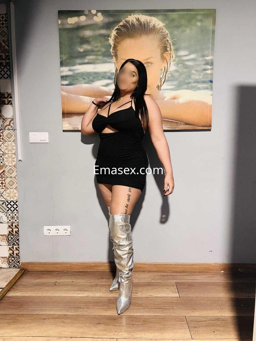 Puta posando en Barcelona con un vestido negro y botas plateadas, frente a una pared decorada.