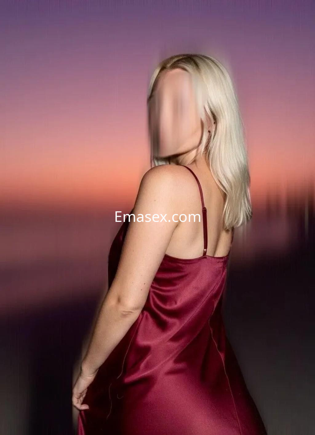Escort con cabello rubio y liso, vistiendo un vestido de seda burdeos, de pie frente a un atardecer en Coruña.