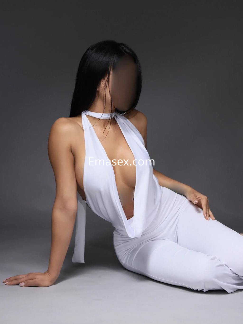 Escort en Madrid posando sentada, viste un ajustado mono blanco con un escote pronunciado y el cabello largo y lacio.