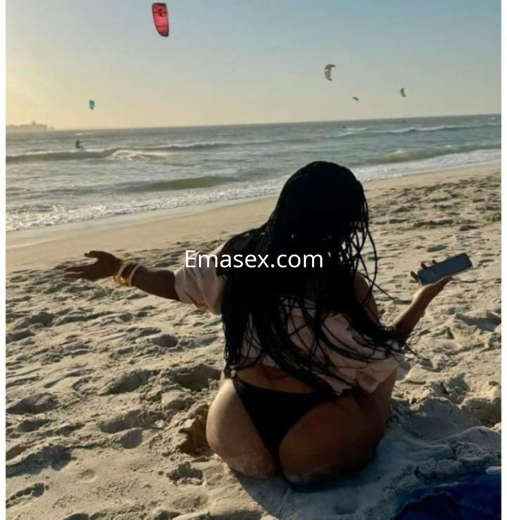 Escort en Ourense sentada en la playa, con cabello largo y suelto, usando un bikini negro y disfrutando del paisaje.