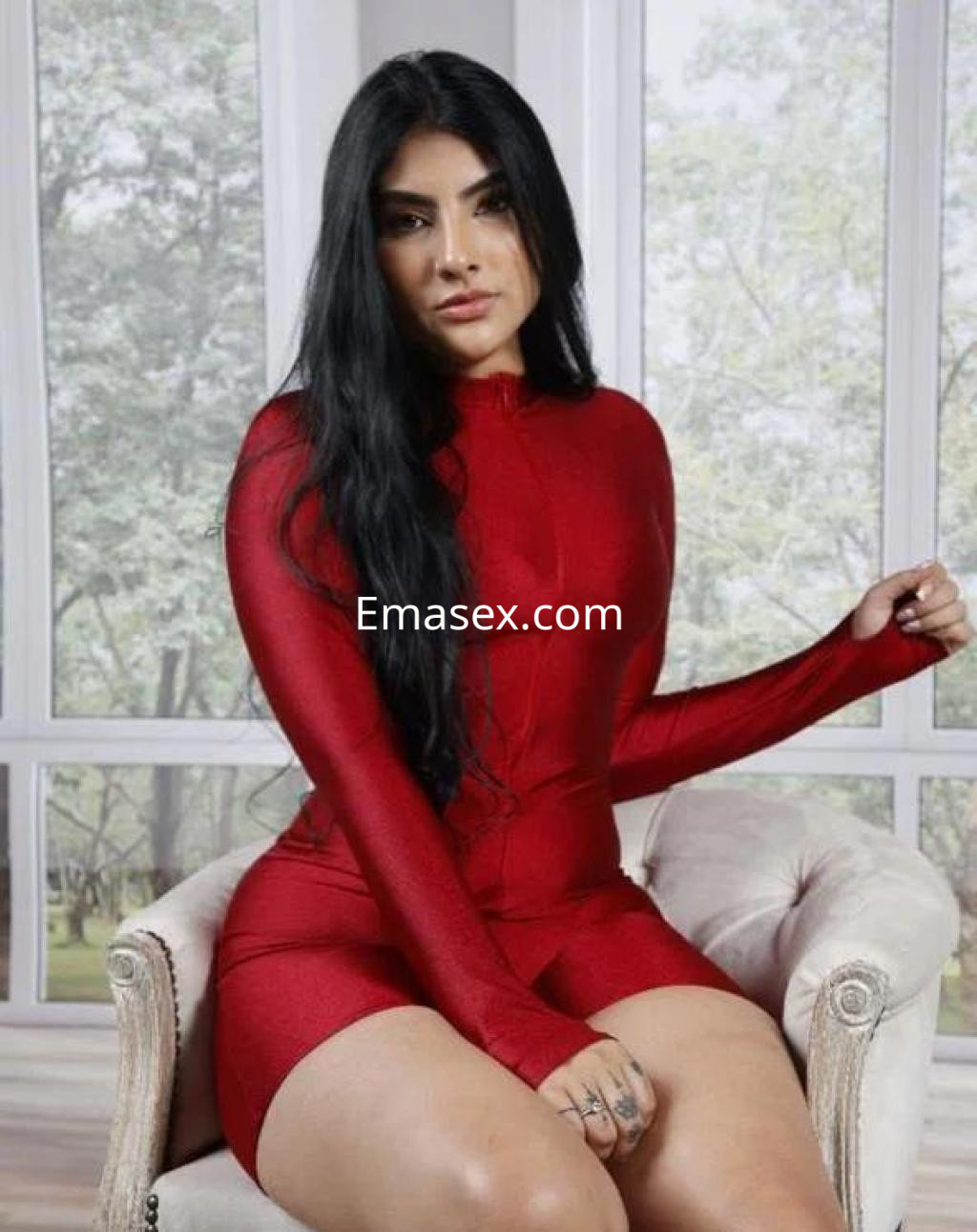 Escort en Tarragona con pelo largo y oscuro, vistiendo un bodysuit rojo ajustado, sentada en un sillón.