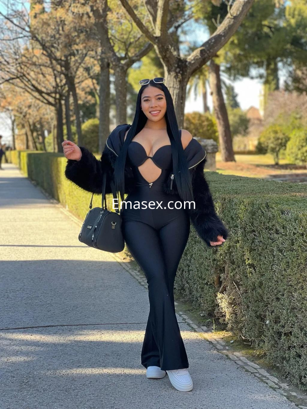 Escort en Zaragoza vistiendo un conjunto negro ajustado con detalles transparentes y un abrigo de pelaje, sonriente en un parque.