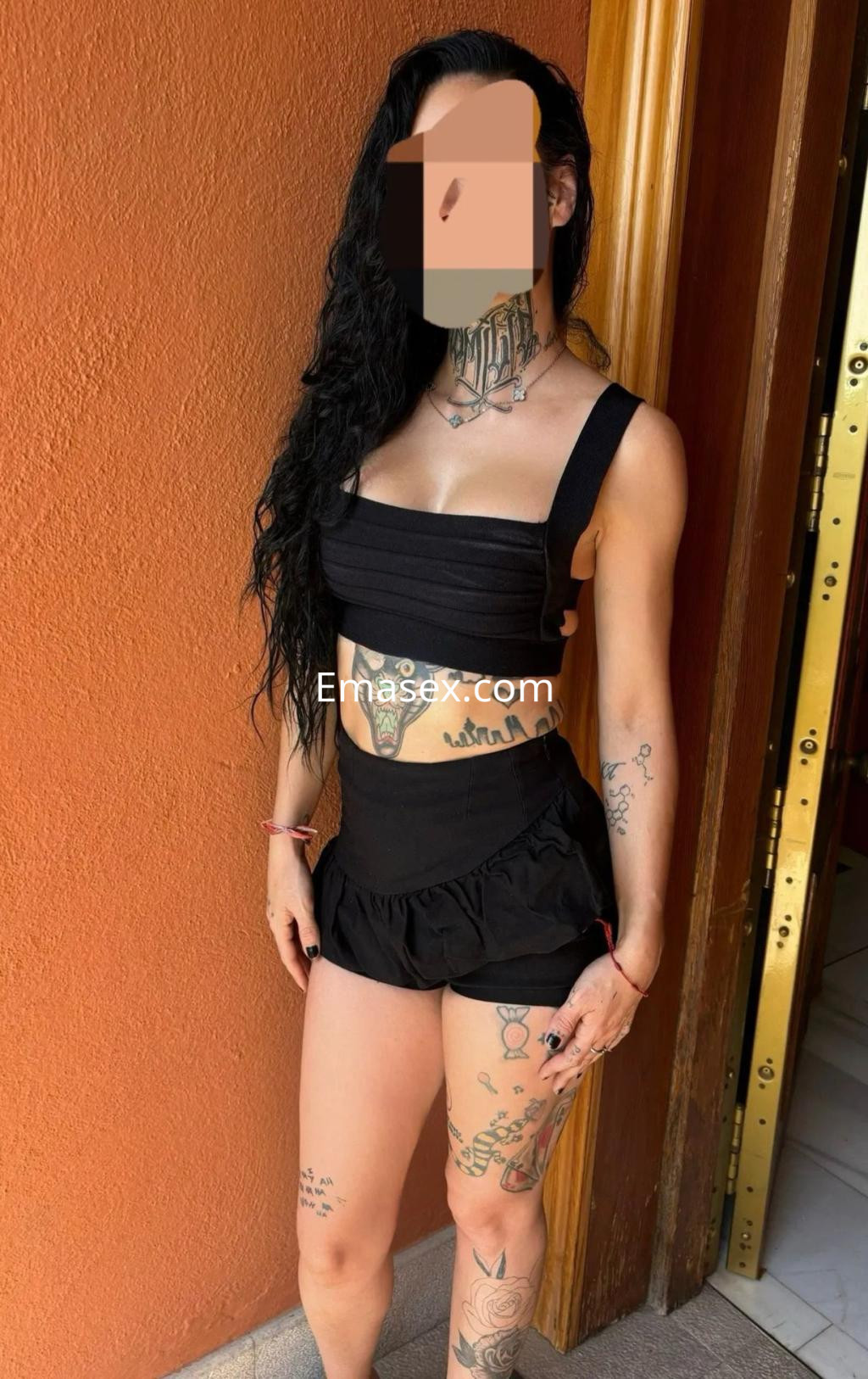 Escort en Málaga con cabello largo y rizado, luciendo un conjunto negro de top y short, mostrando tatuajes en los brazos.