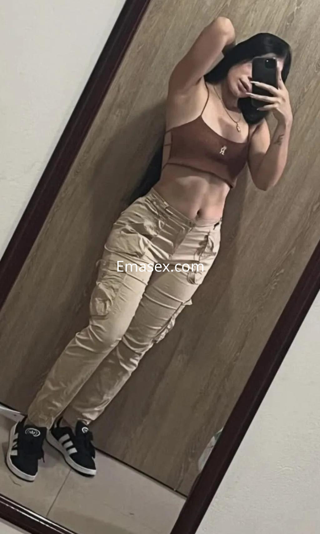 Escort en Málaga posando frente a un espejo con un top marrón y pantalones cargo beige, mostrando su figura.