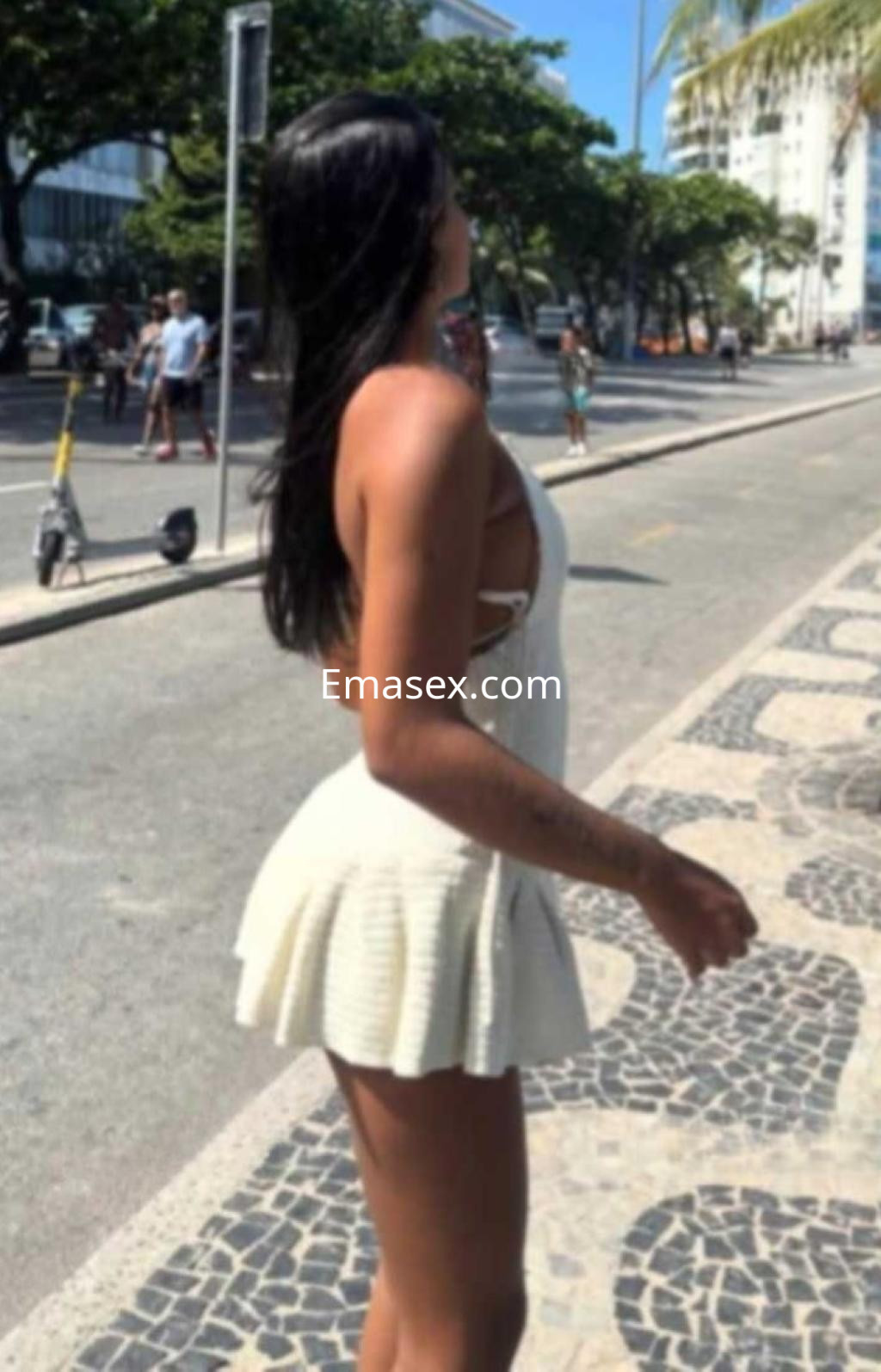 Puta en Alicante con un vestido blanco corto y escote, de pie en una calle soleada, mirando al horizonte.
