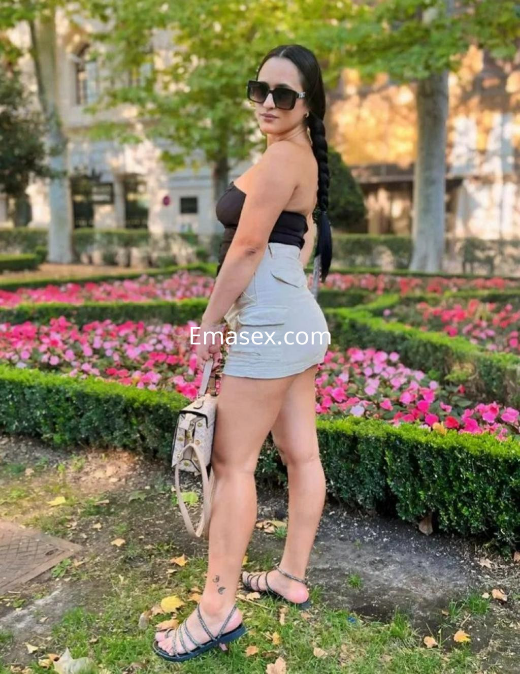 Puta de cabello largo trenzado, viste un top negro y una falda corta, posando en un parque de Madrid.