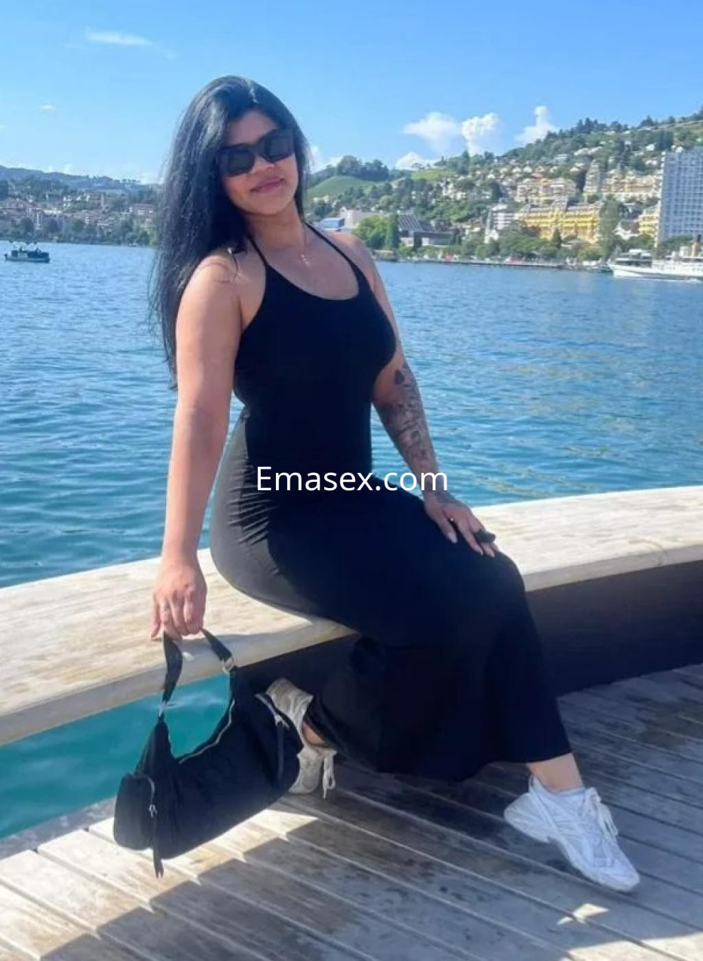 La escort en Madrid luce un vestido negro sin mangas, sentada junto al agua, con cabello largo y oscuro.