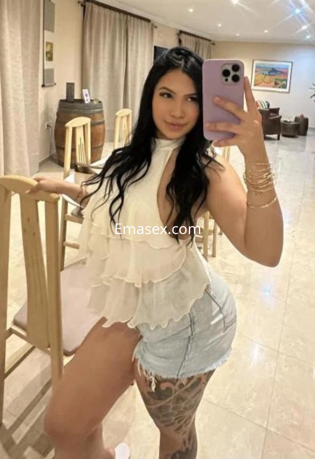 Escort en Cádiz posando con blusa blanca y shorts de mezclilla, mostrando un tatuaje en la pierna.
