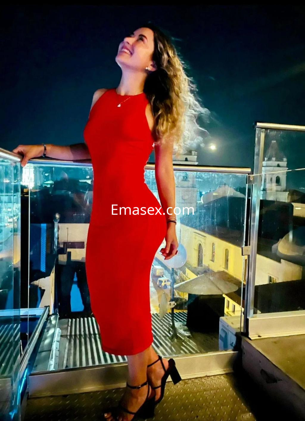 La puta sonríe mientras posa en un balcón con una vista nocturna, luciendo un vestido rojo y tacones.
