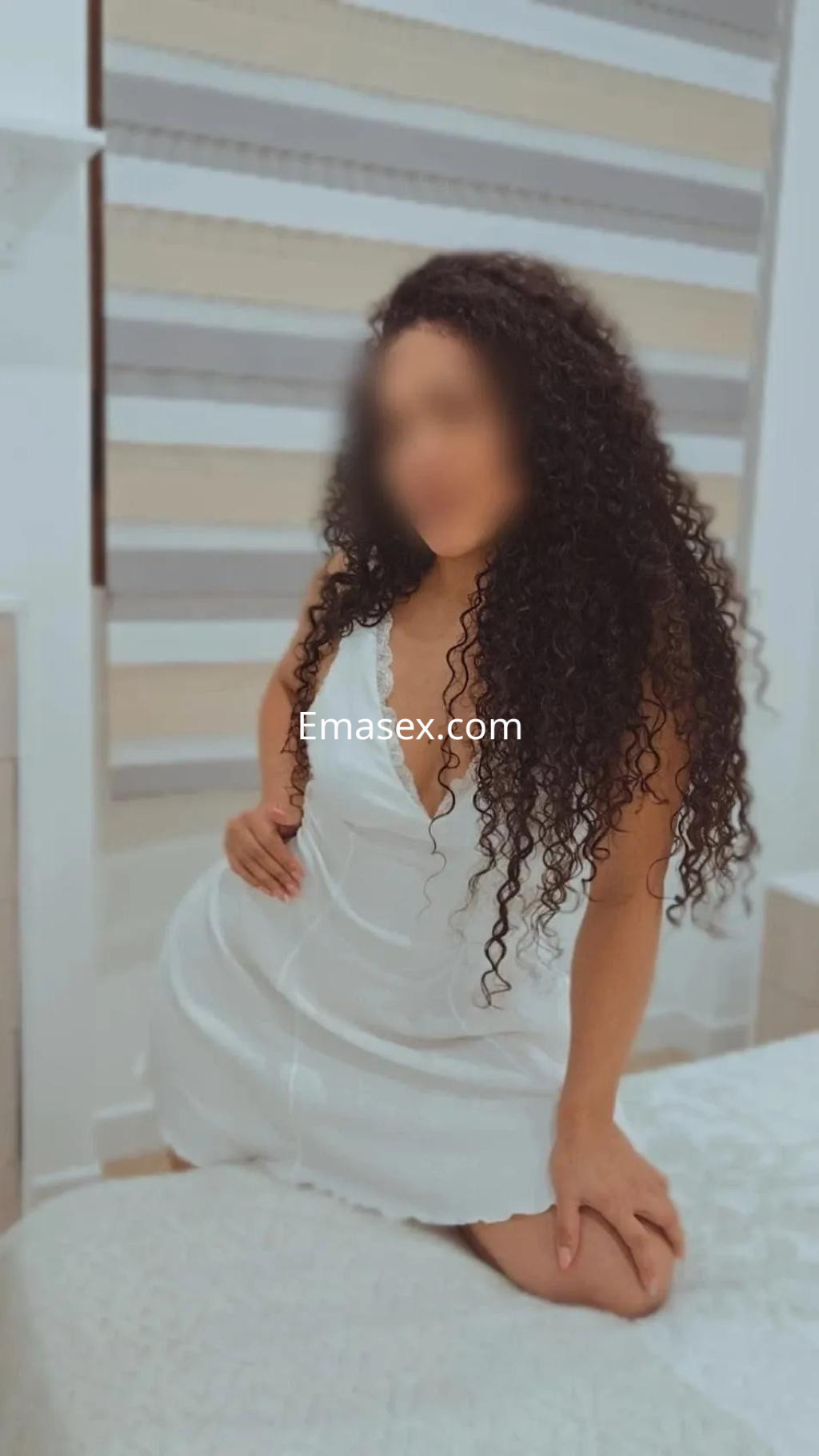 La puta está sentada sobre una cama en Córdoba, luciendo un vestido blanco y con el cabello rizado y oscuro.