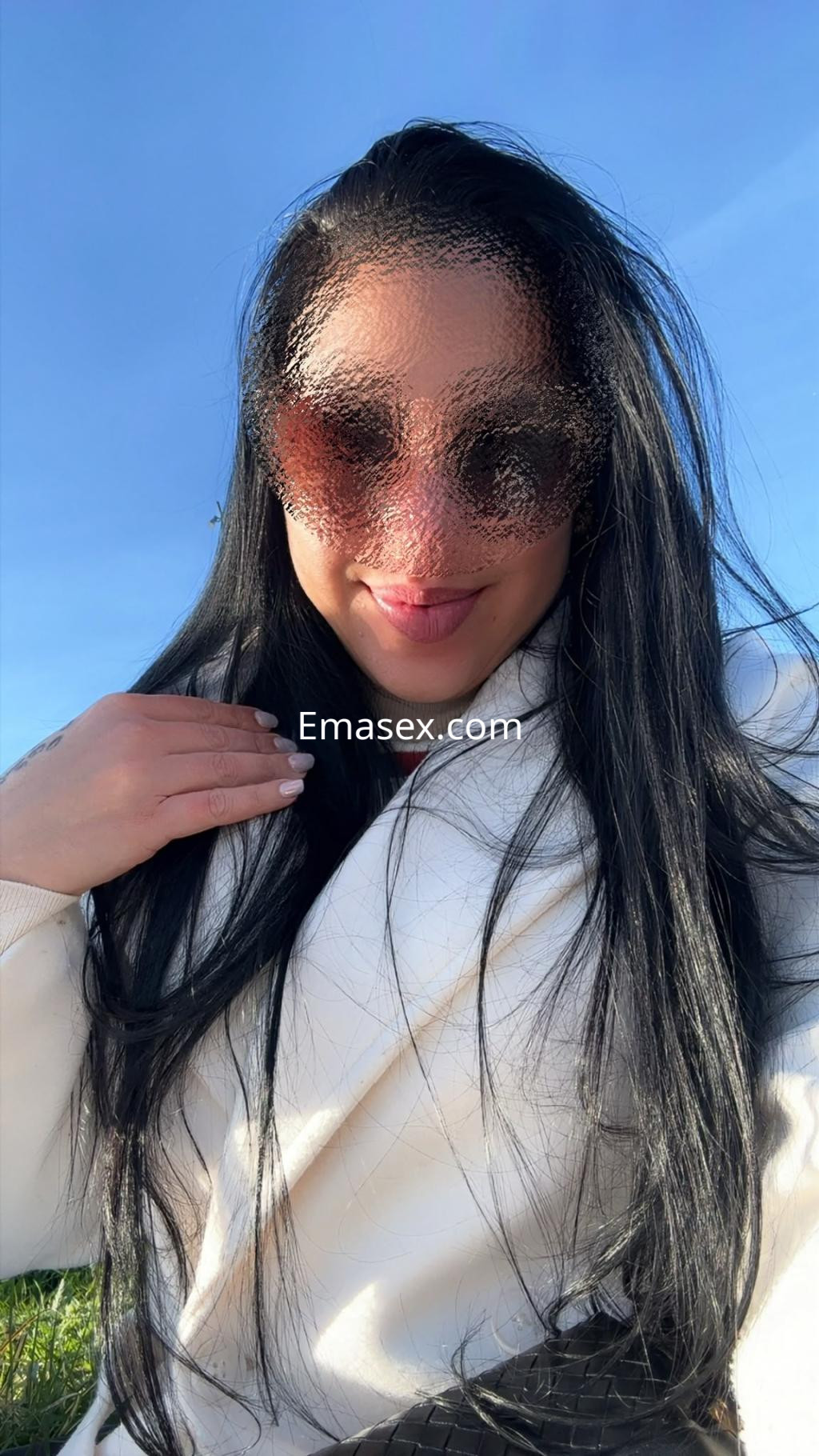 Escort en Vigo con cabello largo y lacio, usando gafas de sol y abrigo blanco, sonríe en un entorno al aire libre.