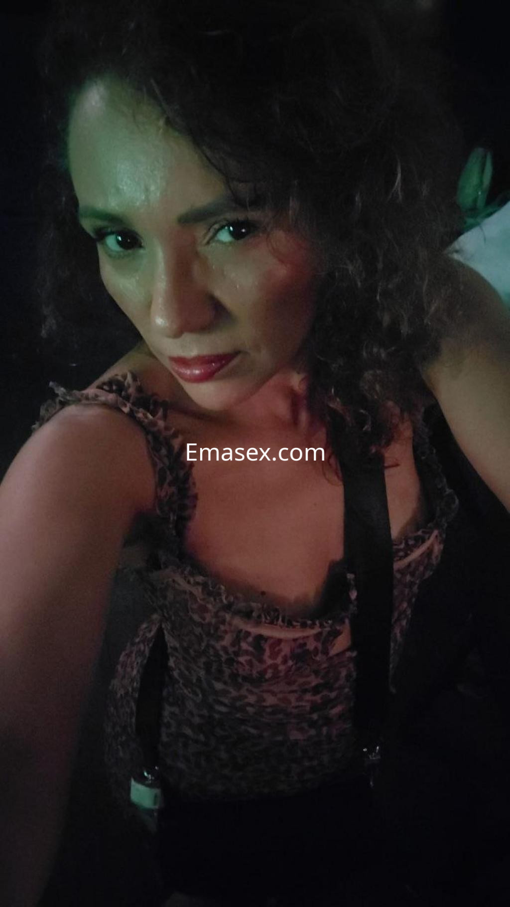 Escort en Santander, con cabello rizado y top de estampado animal, posando con una mirada seductora en un entorno oscuro.