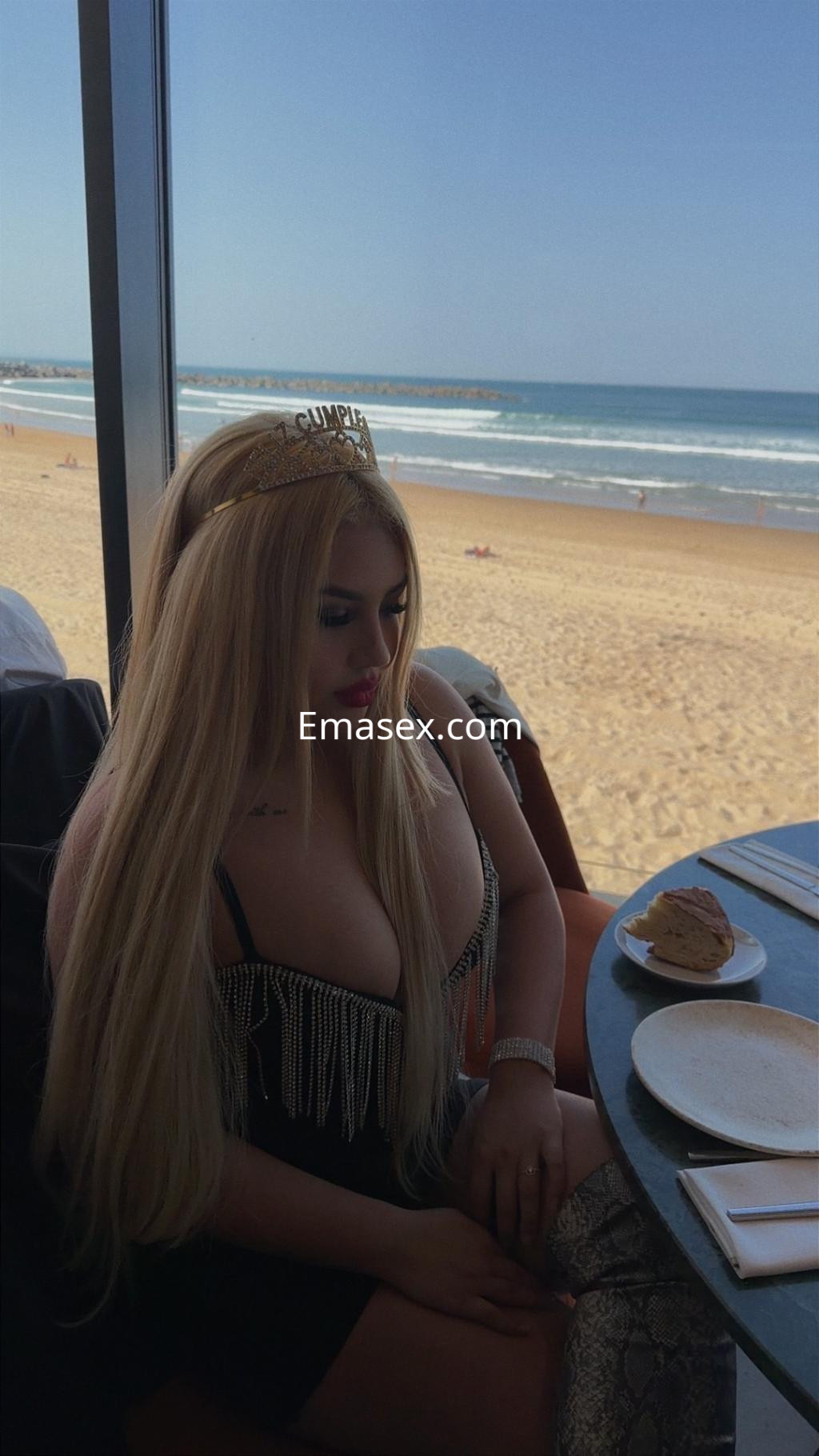 Puta en Donosti con vestido negro con flecos, cabello largo y rubio, sentada junto a una ventana con vista a la playa.