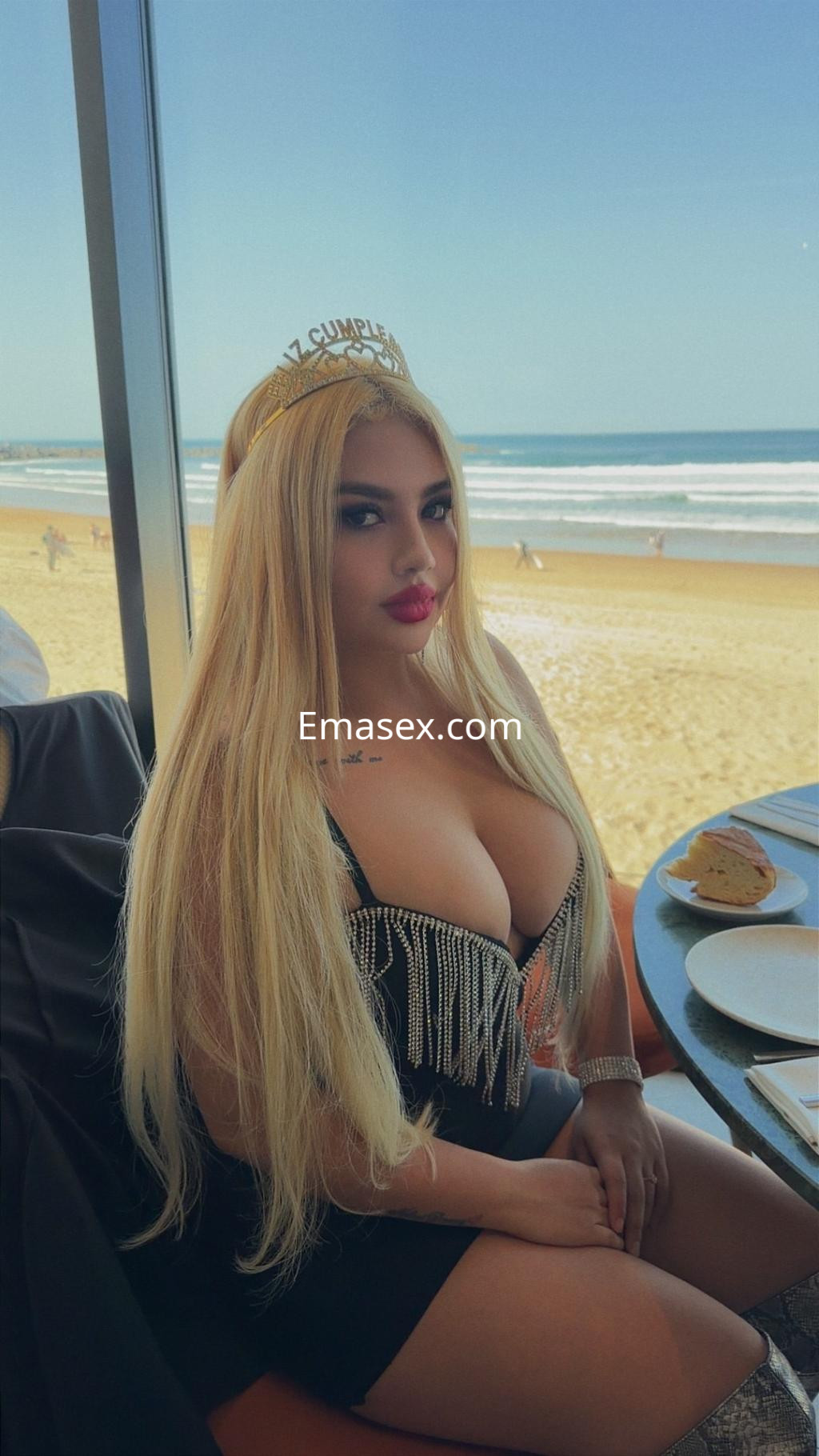 Puta en Donosti con vestido negro ajustado y cabello largo y rubio, sentada en un restaurante con vista a la playa.
