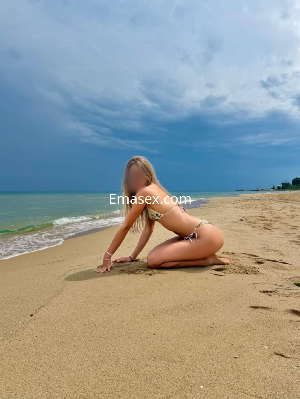 Escort en Girona, posando de rodillas en la playa con un bikini blanco, frente a un mar tranquilo y cielo nublado.