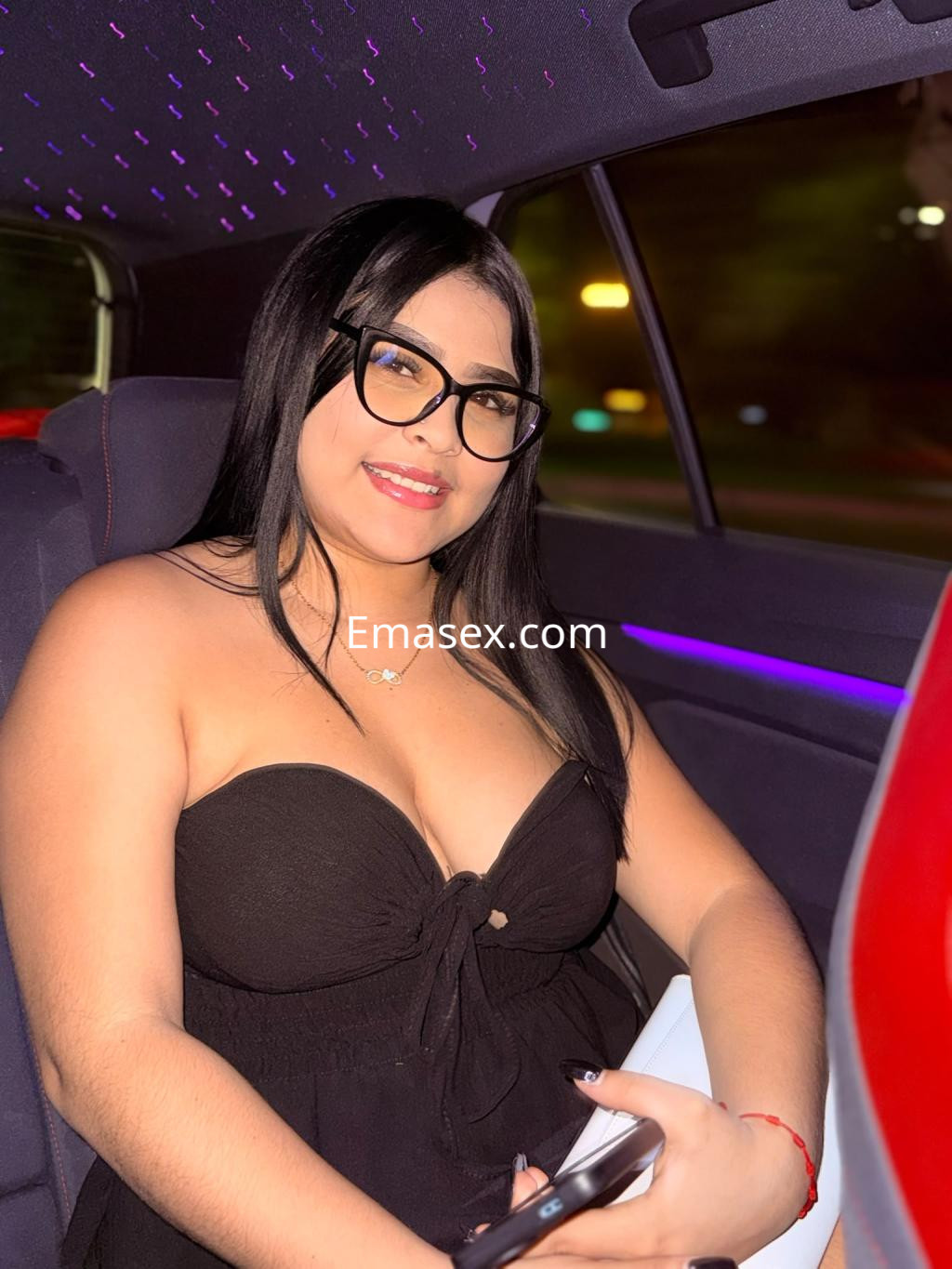 Puta en Alicante con un vestido negro sin mangas y gafas de pasta, sonriendo mientras está sentada en un coche.
