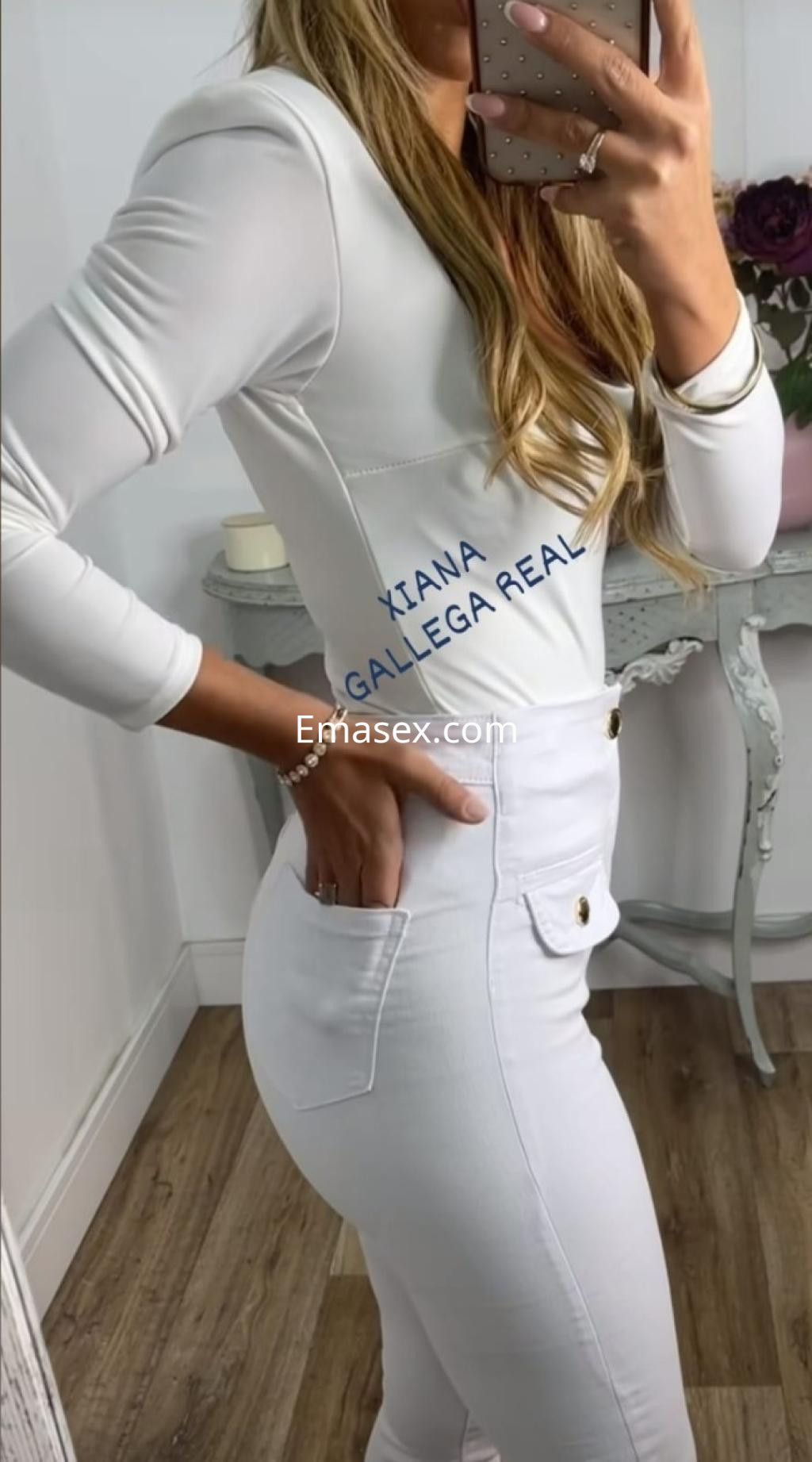 La escort en Santiago viste una blusa blanca ajustada y pantalones blancos, posando de lado frente al espejo.