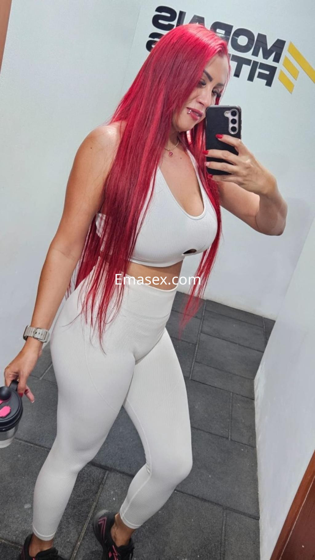 Escort en Gijón con cabello largo y rojo, vistiendo un conjunto deportivo blanco, posando en un gimnasio.