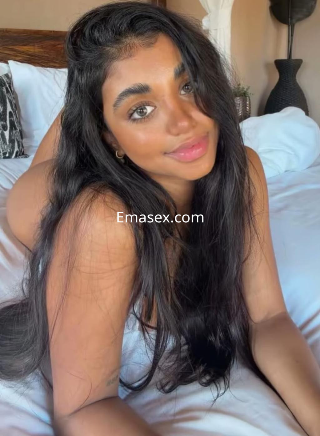 Escort en Tarragona con cabello largo y oscuro, posando en una cama con sábanas blancas y un ambiente acogedor.