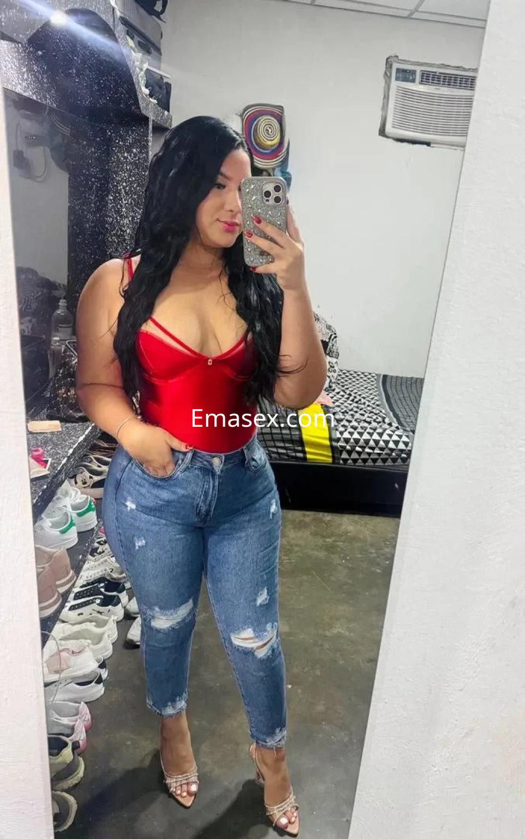 La puta en Alicante lleva un body rojo ajustado y jeans rotos, posando con confianza frente al espejo.
