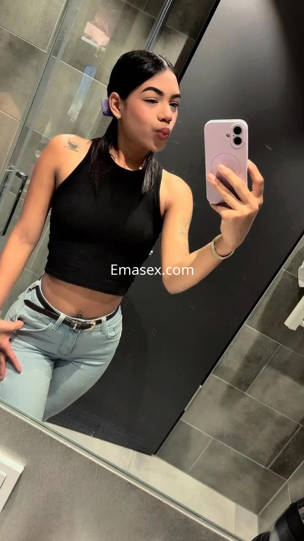 Escort en Alicante, luciendo un top negro y jeans claros, posando frente al espejo con un peinado elegante.