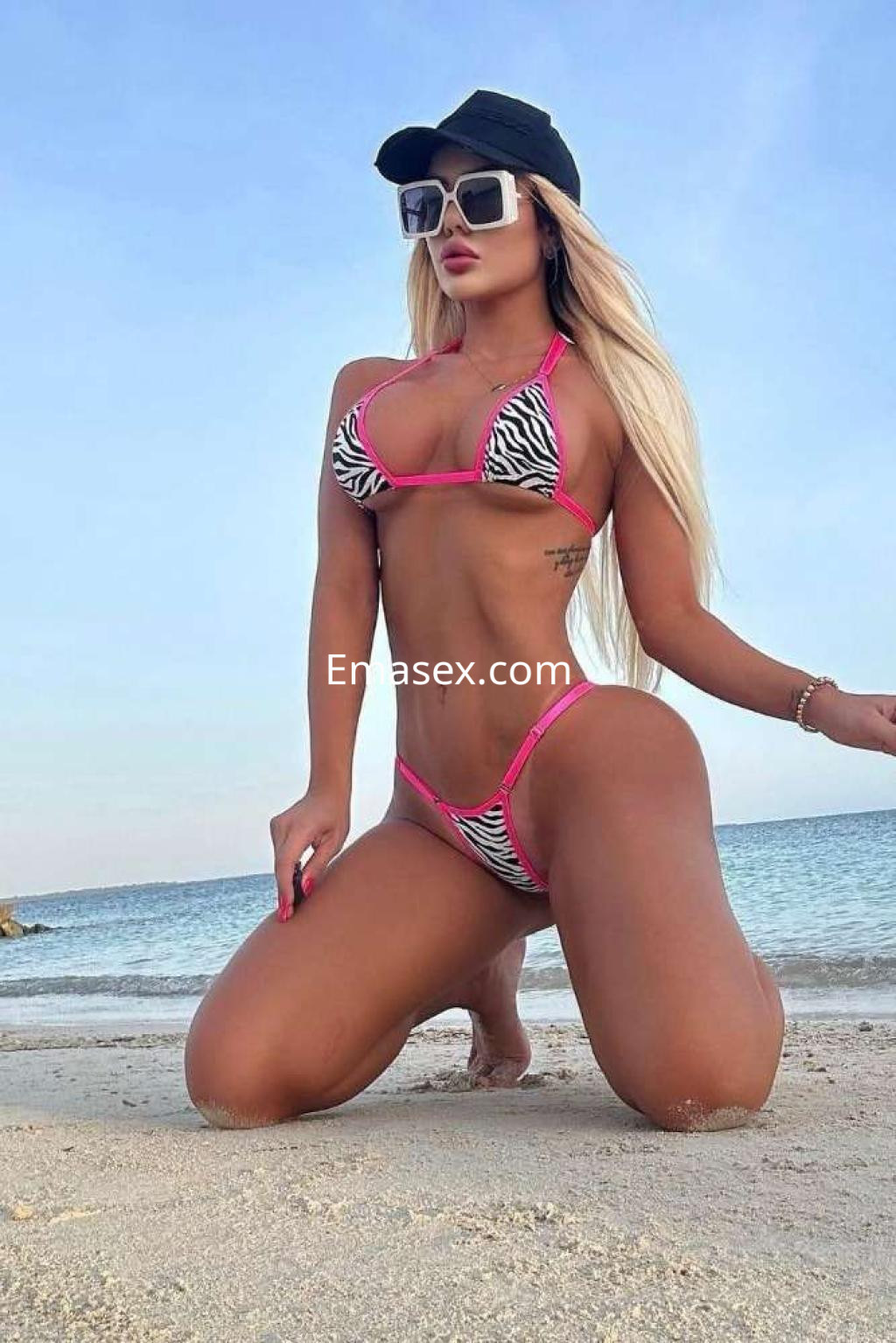 Escort en Ibiza luciendo un bikini de cebra rosa y negro, con gorra y gafas de sol, posando en la playa.