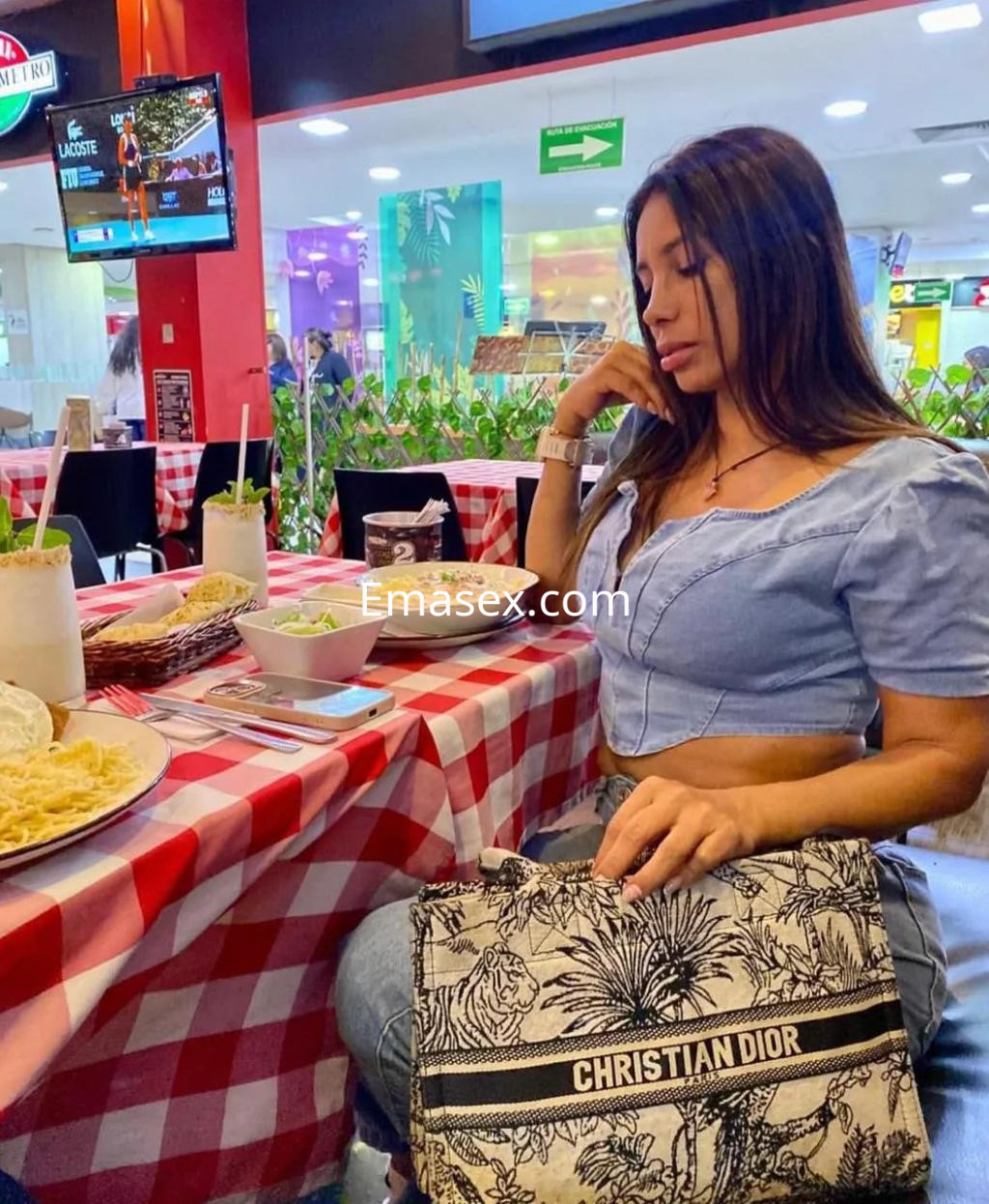 Escort en Palma de Mallorca sentada en una mesa con mantel de cuadros, luciendo blusa azul y sosteniendo un bolso.