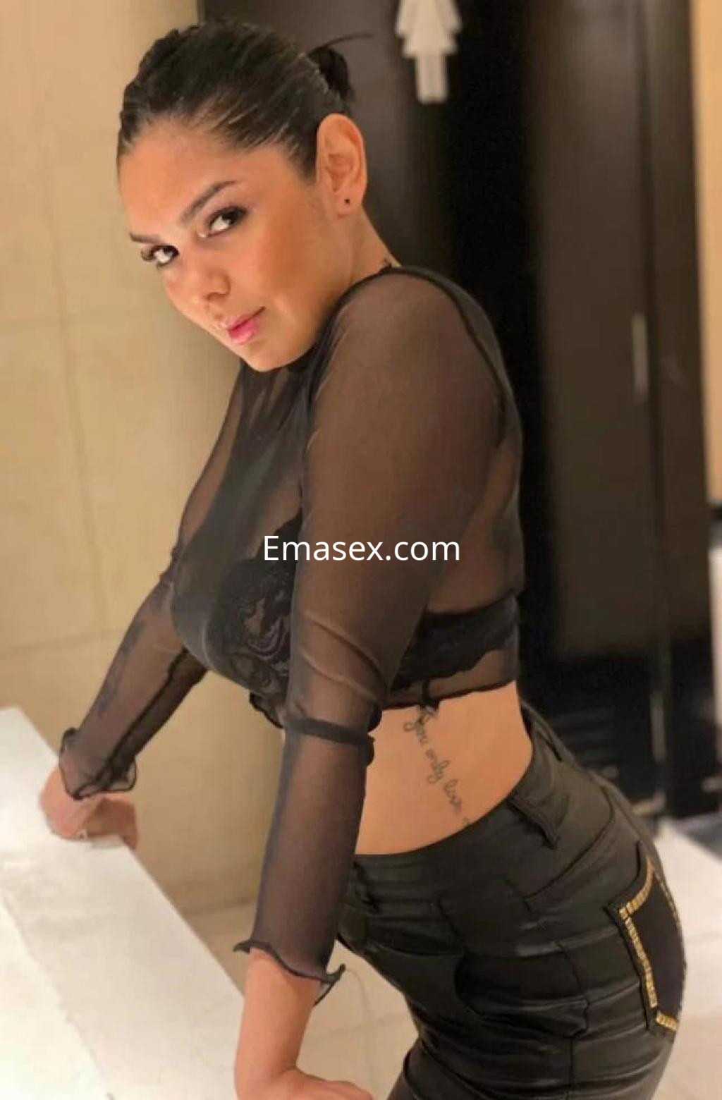 Escort en Palma de Mallorca con blusa transparente y pantalones ajustados, posando en un entorno elegante.