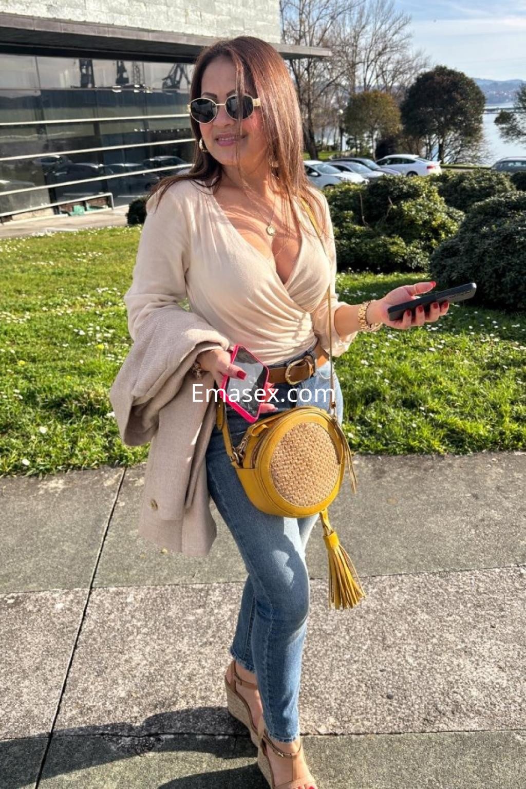 Escort en Coruña, lleva una blusa clara y jeans ajustados, con un bolso amarillo y gafas de sol, sonriendo al caminar.