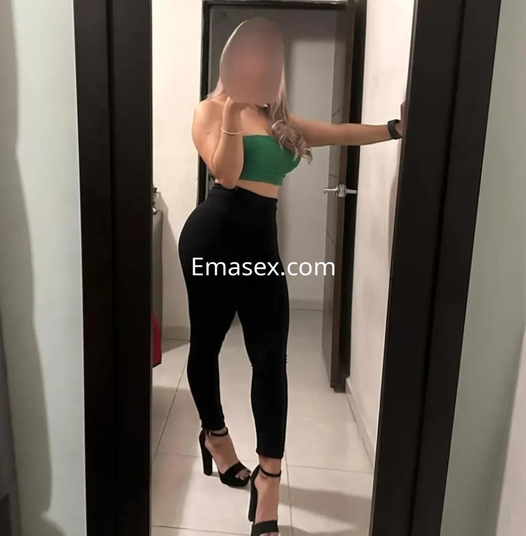 Puta en Murcia posa frente al espejo con top verde y pantalones negros, luciendo tacones altos.