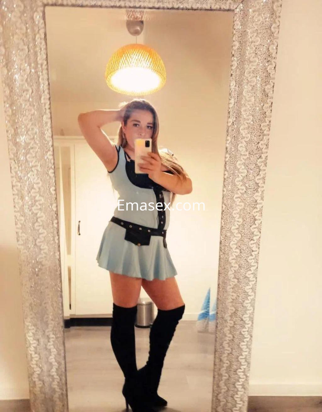 Escort en Pontevedra posando frente a un espejo con un vestido corto azul y botas altas, mostrando su cabello largo.