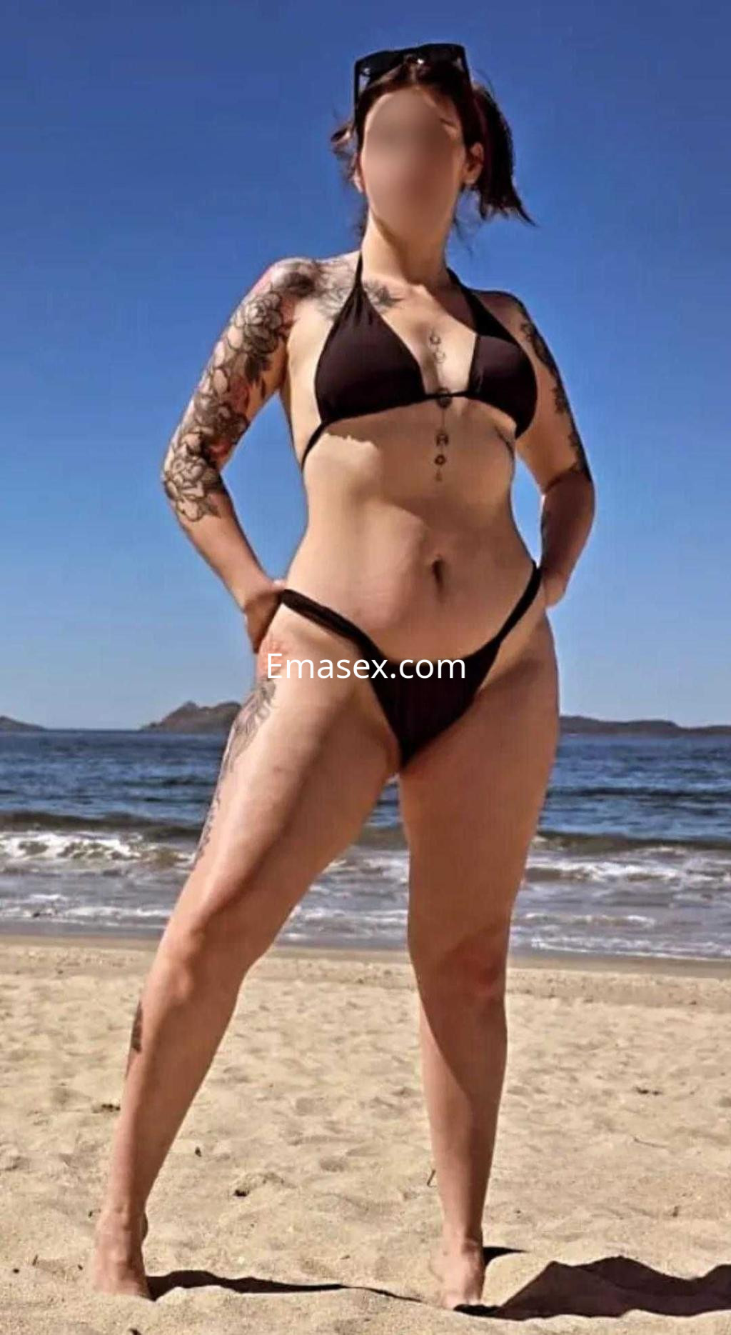 Escort en Vigo posando en la playa con bikini negro, luciendo tatuajes y gafas de sol, bajo un cielo despejado.