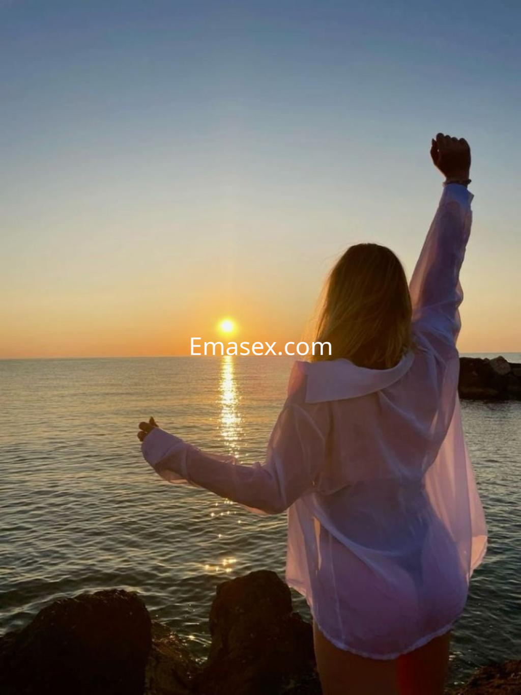 Escort en Tenerife de espaldas, con una blusa blanca y brazos alzados, disfrutando de la puesta de sol sobre el mar.
