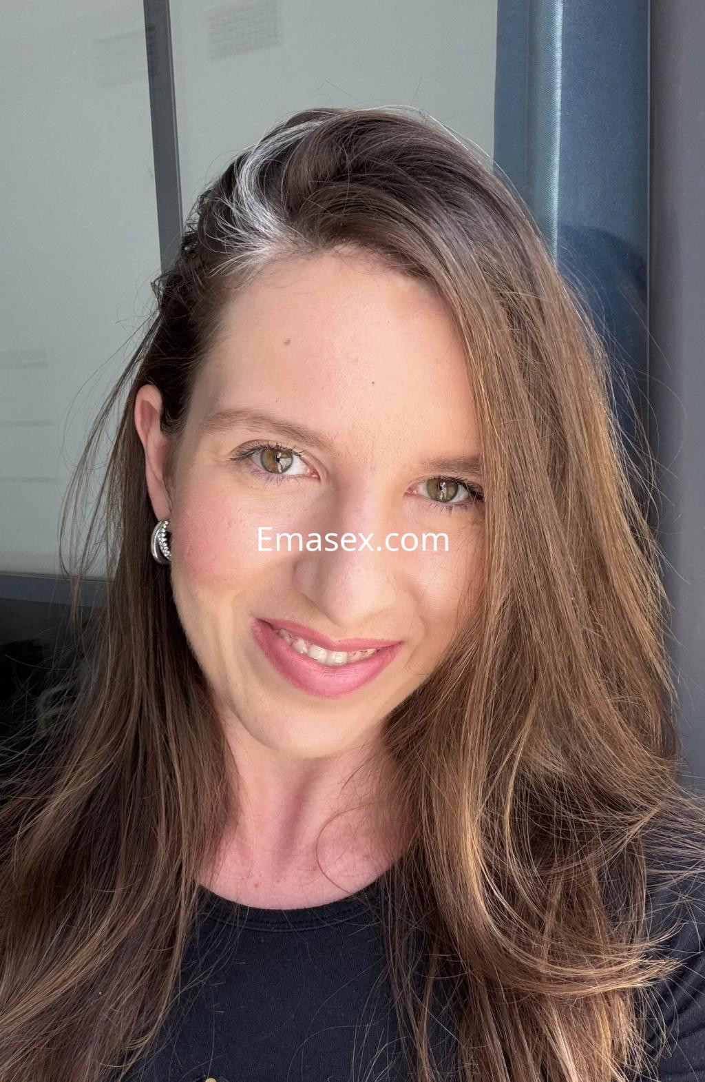 Escort en Málaga con cabello largo y suelto, sonrisa amigable y pendientes brillantes frente a una ventana.