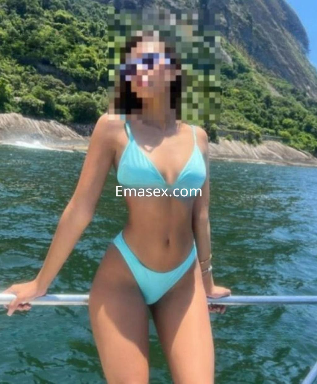 Escort en Zaragoza luciendo un bikini azul claro, de pie en un barco con un fondo natural de paisaje costero.