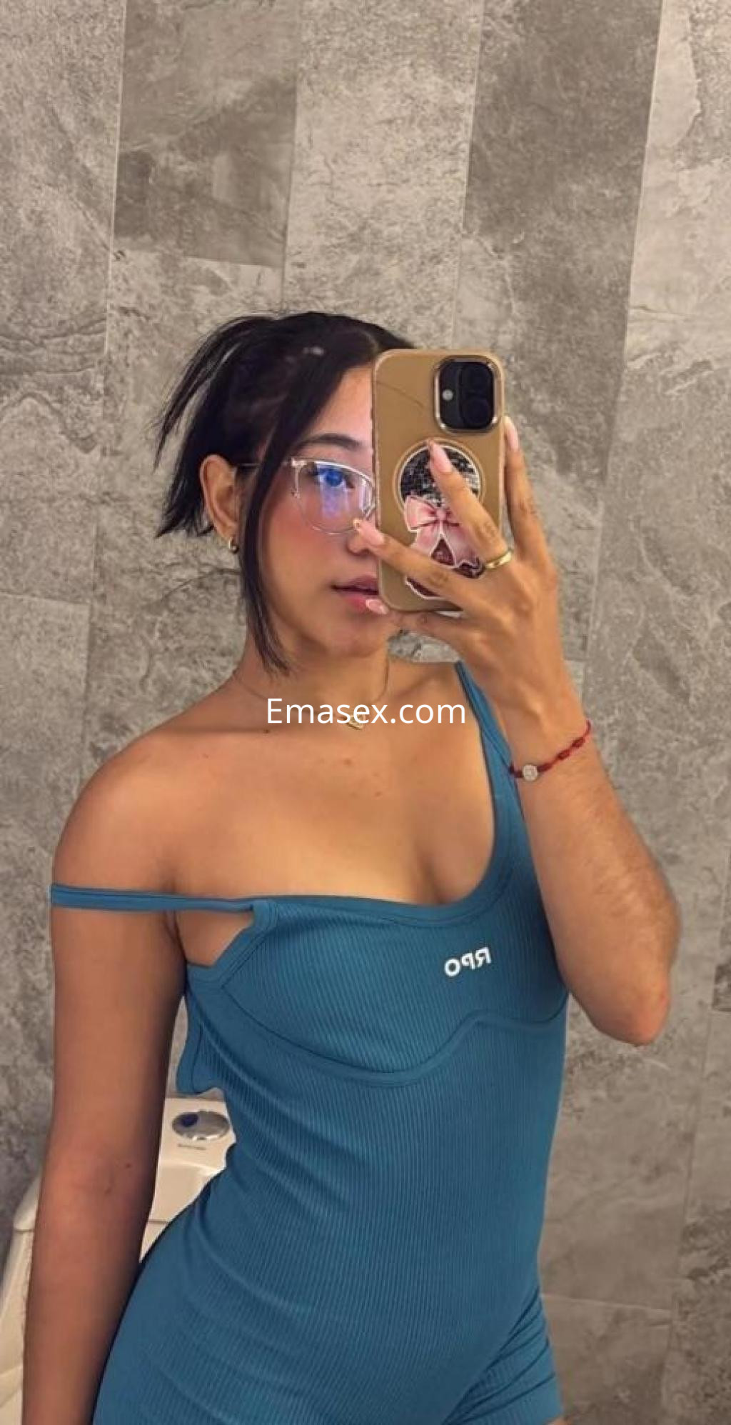 Puta en Coruña, posando con un mono azul y gafas en un baño con azulejos grises.