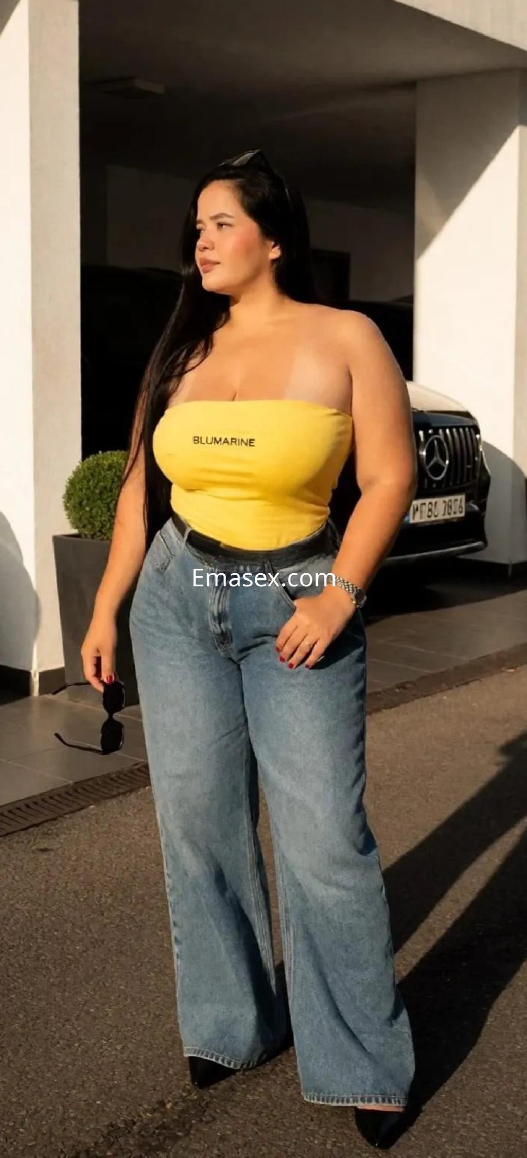 Escort en Vigo posando con una blusa amarilla sin mangas y pantalones de mezclilla anchos, luciendo confiada.