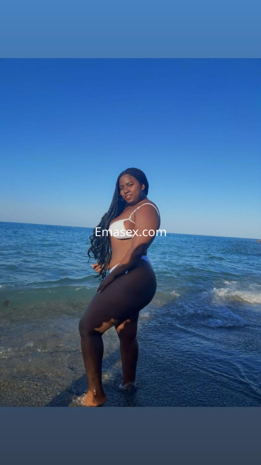 Puta de piel oscura y cabello trenzado, posa en la playa de Granada con un bikini blanco junto al mar.
