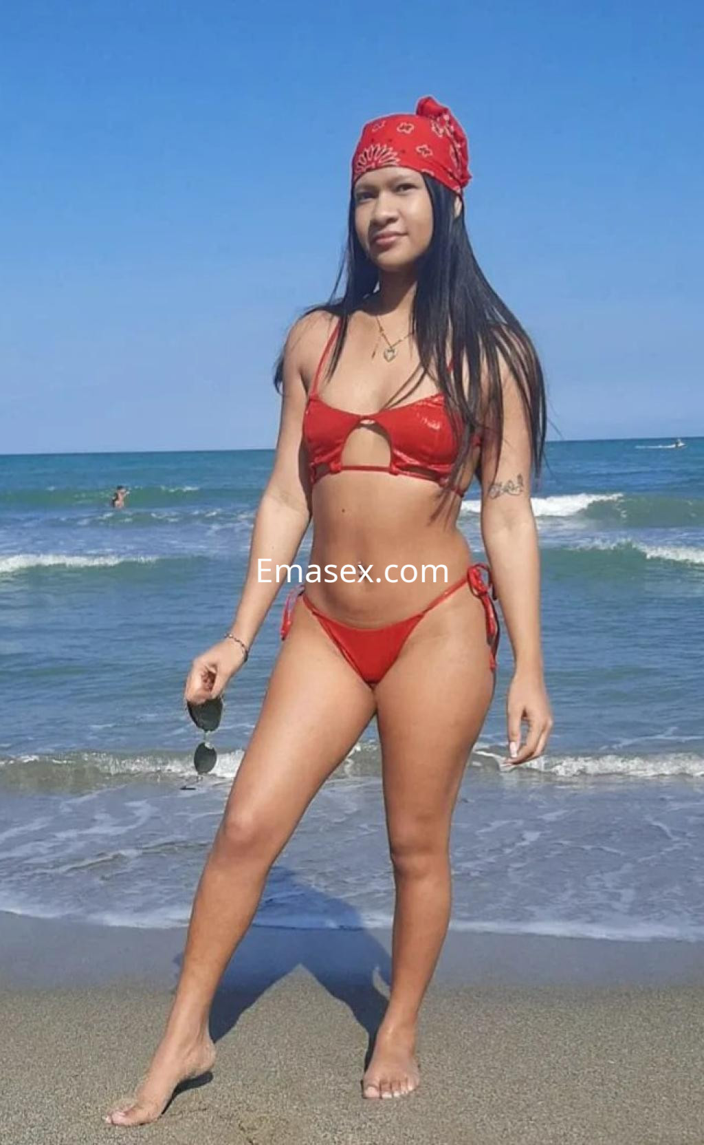 La puta posa en la playa de Zaragoza, luciendo un bikini rojo y un pañuelo en la cabeza bajo un cielo despejado.
