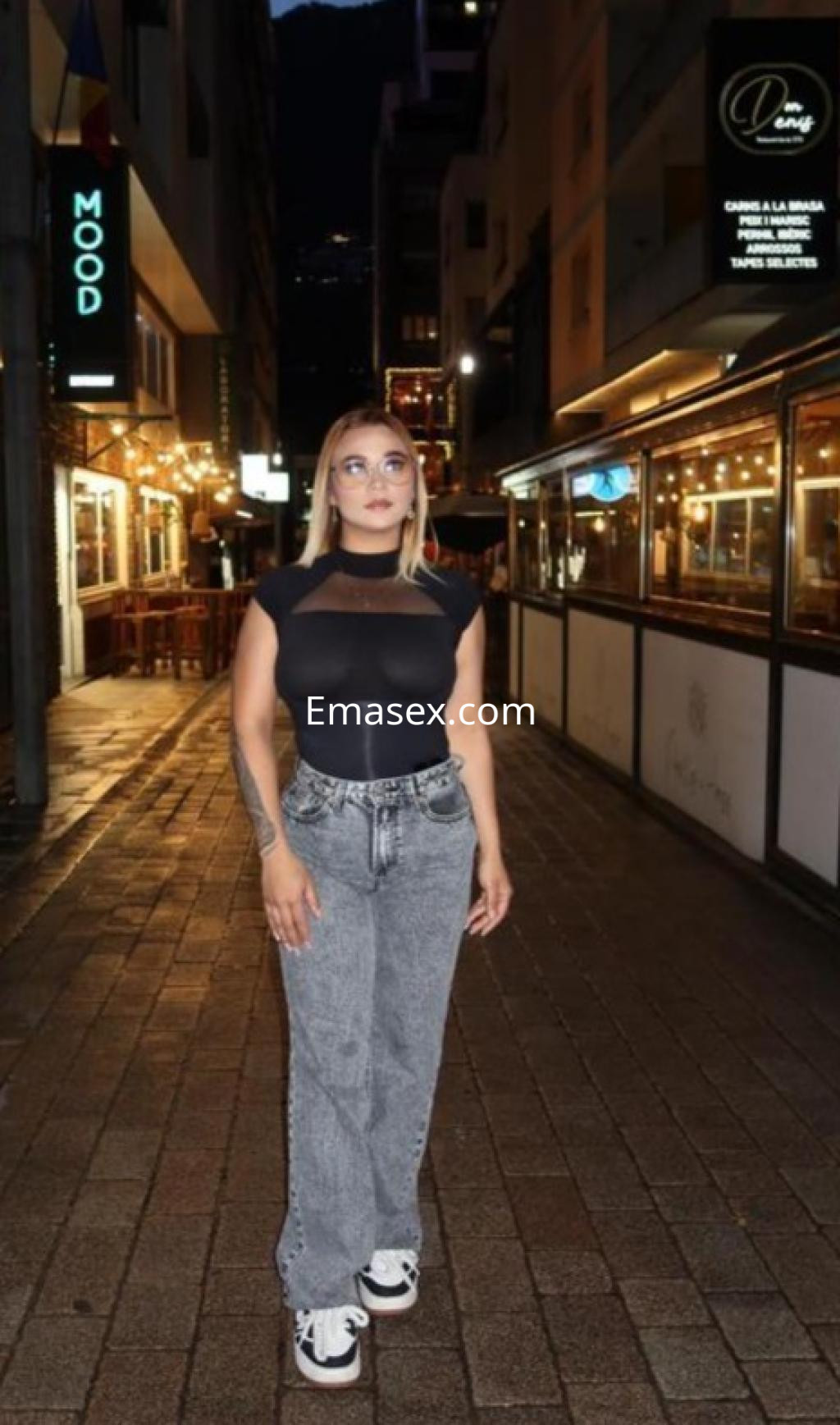 Puta en Lleida viste una blusa negra ajustada y pantalones grises, posando en una calle iluminada por restaurantes.