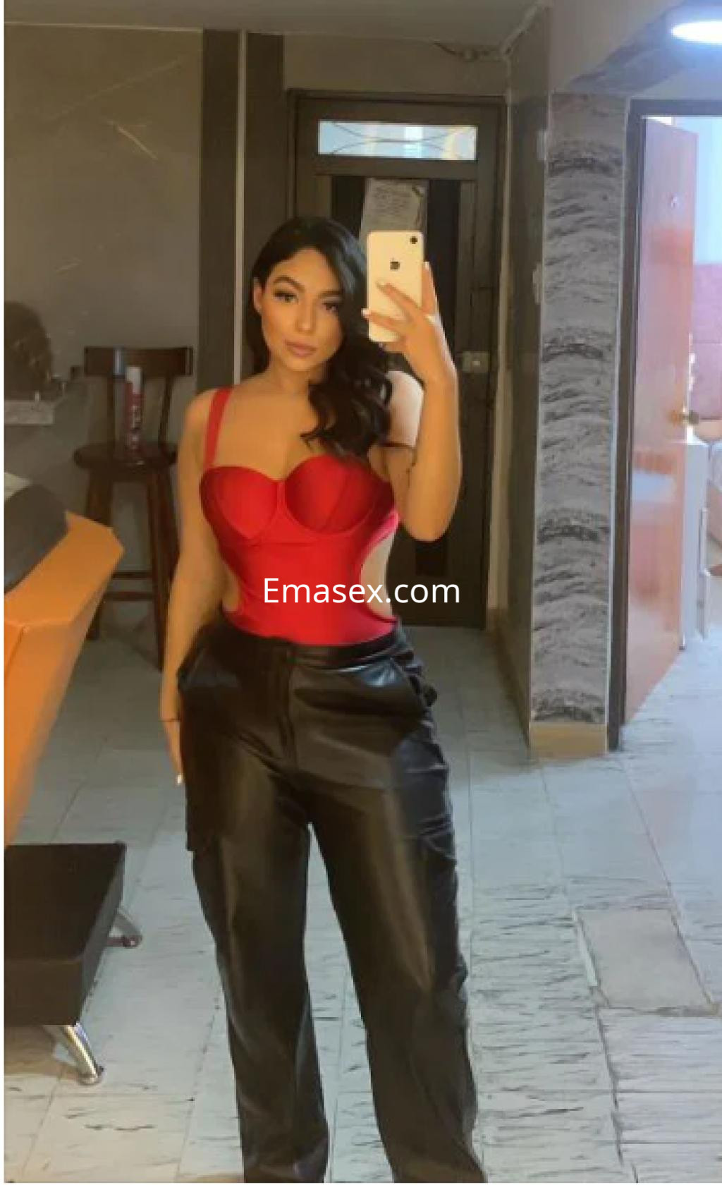Puta en Lleida con un body rojo y pantalones de cuero negros, posando en un ambiente interior moderno.
