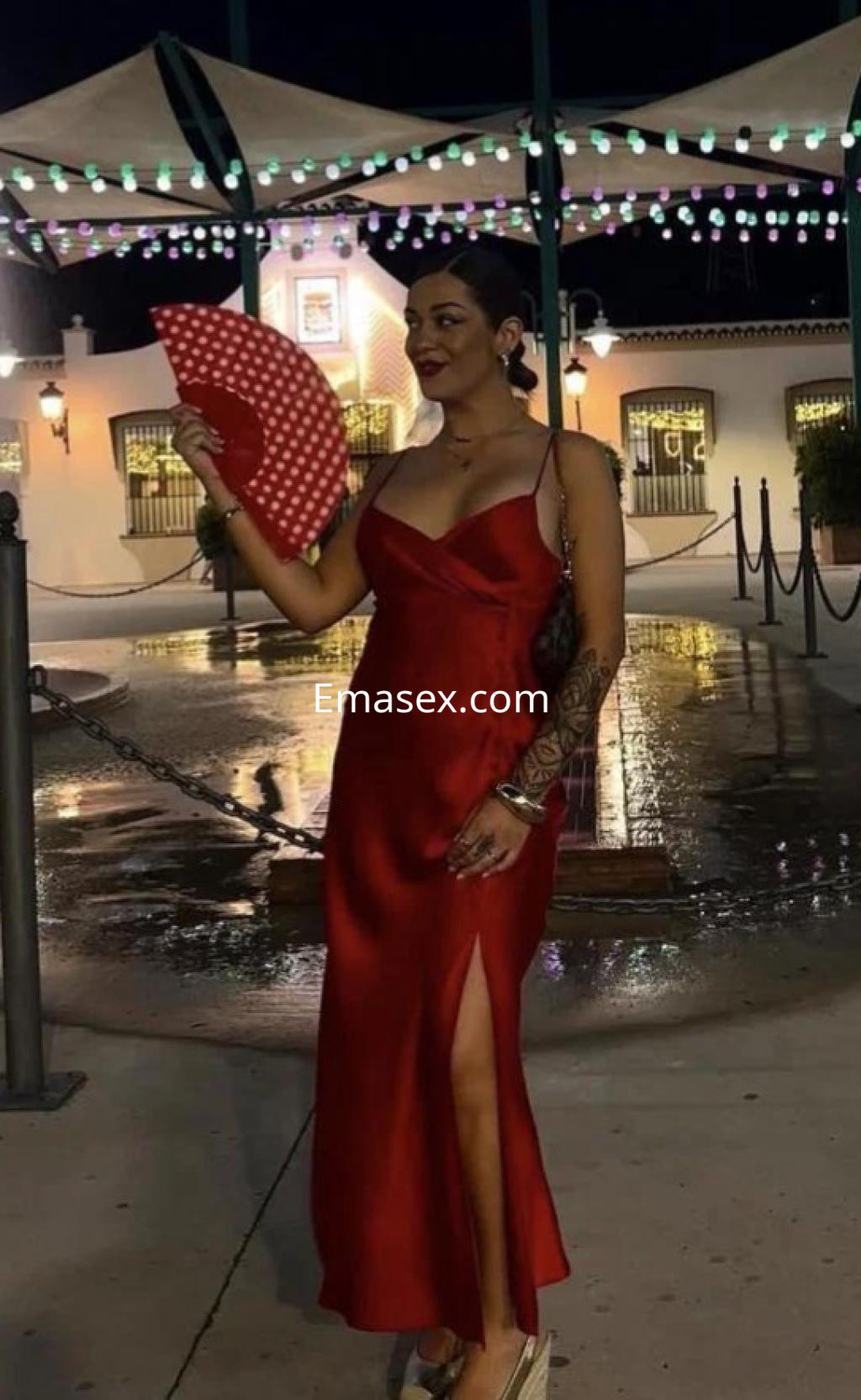 La puta luce un vestido rojo con una abertura lateral, sosteniendo un abanico en un entorno iluminado de Huelva.