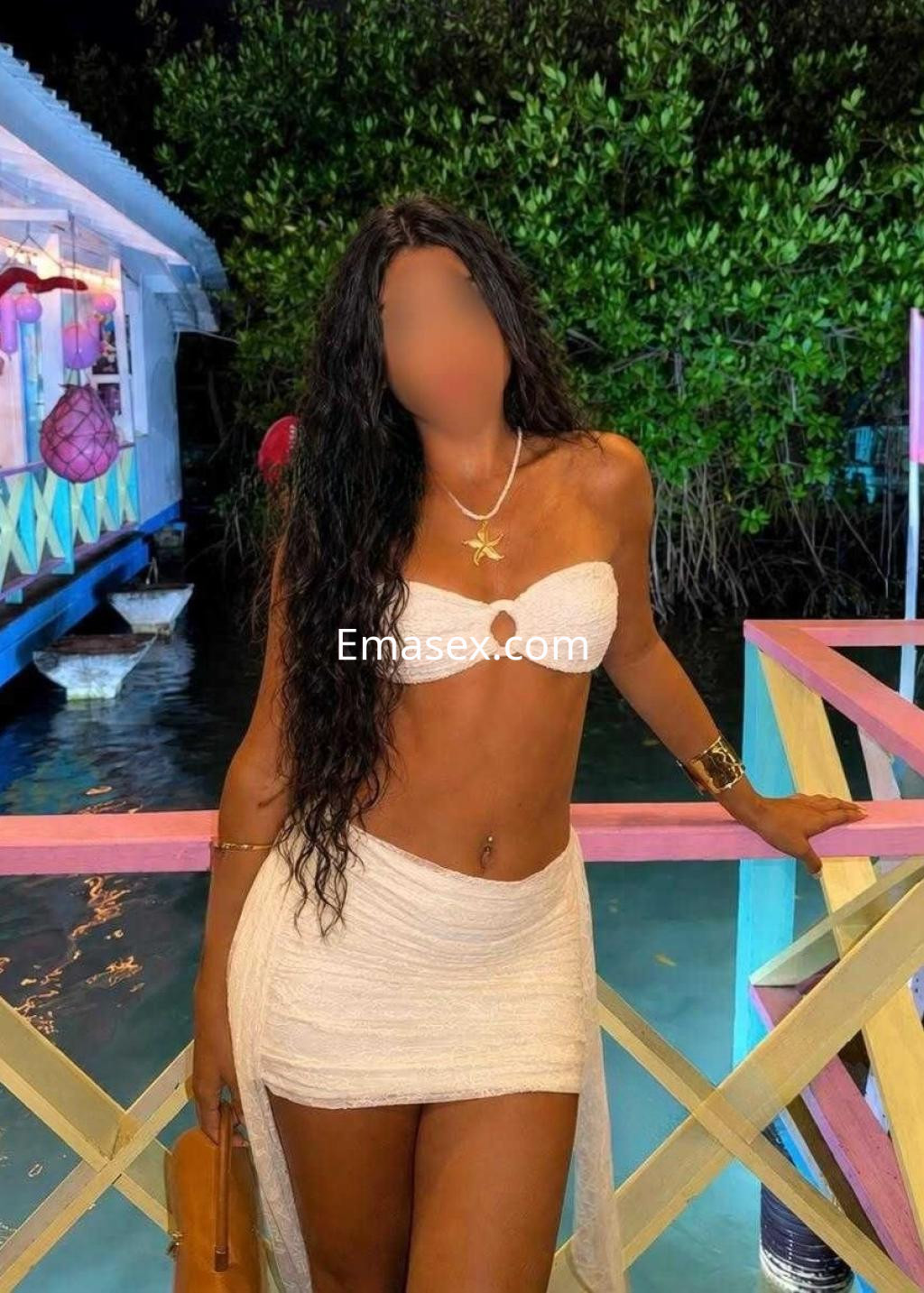 Escort en Huelva con cabello largo y rizado, usando un bikini blanco y una falda corta, posando junto al agua.