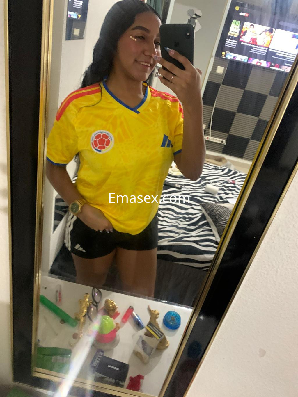 Escort en Granada, posando frente al espejo con camiseta amarilla de fútbol y pantalones cortos negros.