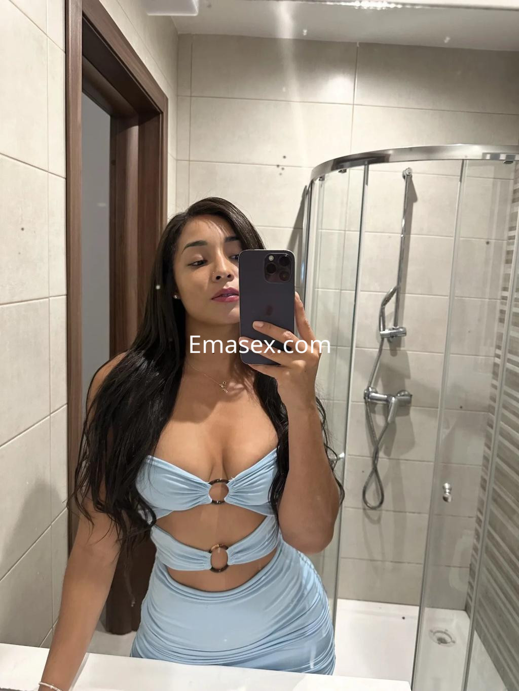 La puta posa en un vestido azul claro con cortes, en un baño moderno de Alicante, mostrando su largo cabello oscuro.