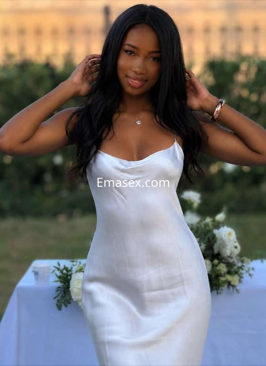 Escort en Ibiza con cabello largo y oscuro, luciendo un vestido blanco ajustado y sonriendo en un ambiente al aire libre.